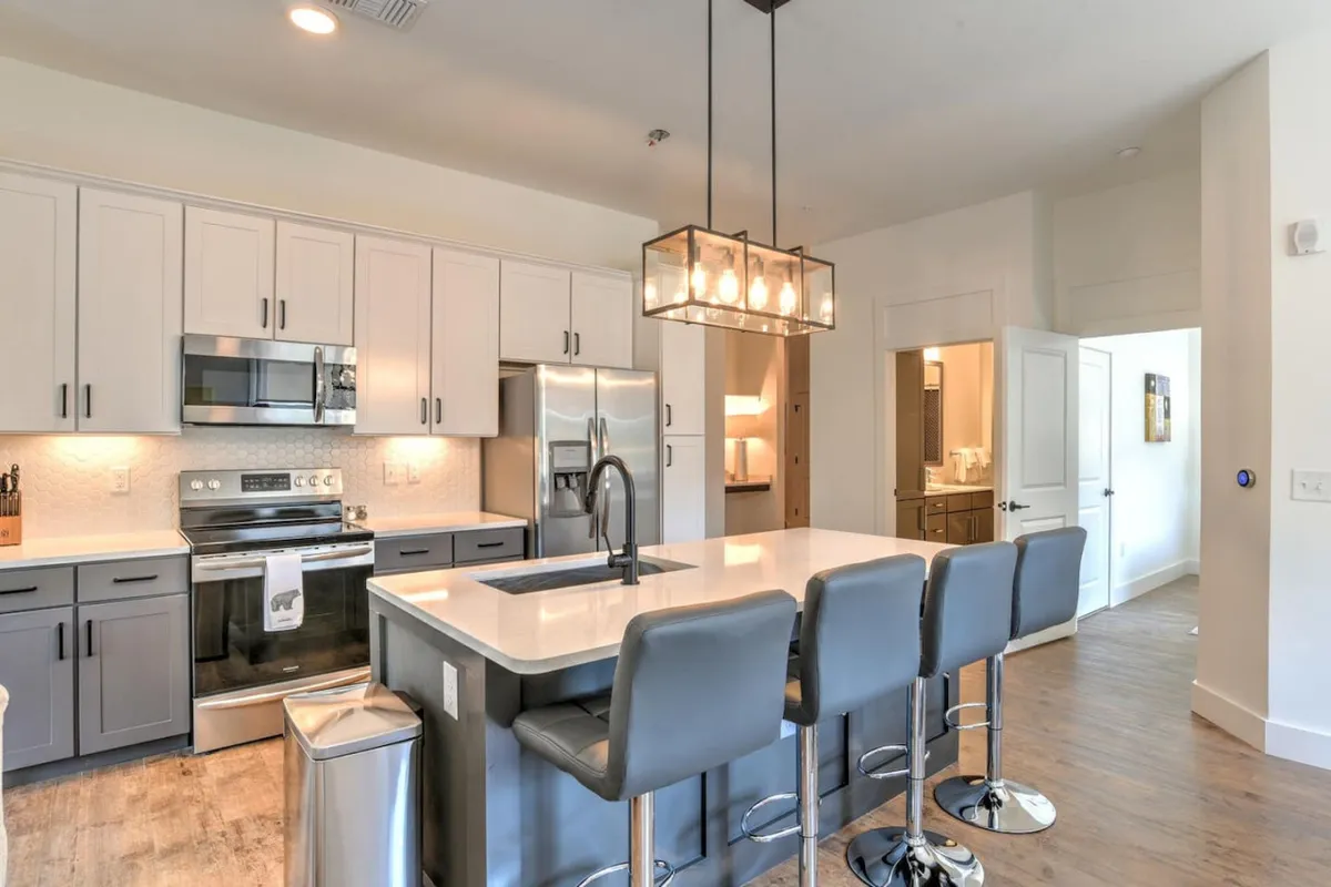 A bright, contemporary kitchen featuring a spacious island with stylish lighting fixtures overhead. Stainless steel appliances line the back wall, and comfortable seating invites guests to gather around. The open space suggests an area perfect for both meal prep and social interaction.