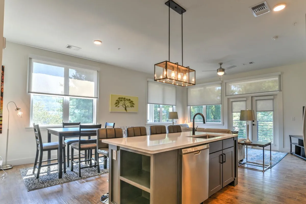 A cheerful, sunlit kitchen and dining area with a large island countertop, dining table, and expansive windows letting in natural light. Elegant decorative lighting adds warmth, and the setting is ready for morning coffee or a memorable dinner party.