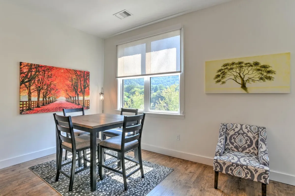 A cozy dining space with a decorative chair, colorful artwork, and a window view that hints at the changing seasons and urban landscape beyond. The space invites guests to enjoy meals, conversation, and the sights of the city.
