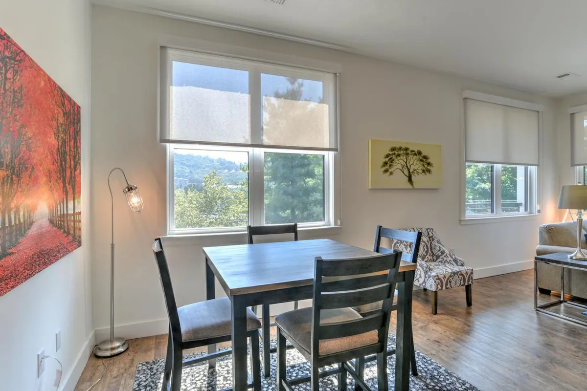 A dining table is situated next to a large window that offers a partial view to the outside, with tasteful artwork on the wall and a relaxed, welcoming atmosphere. The natural light coming through the window hints at the vibrant city life just beyond the glass.