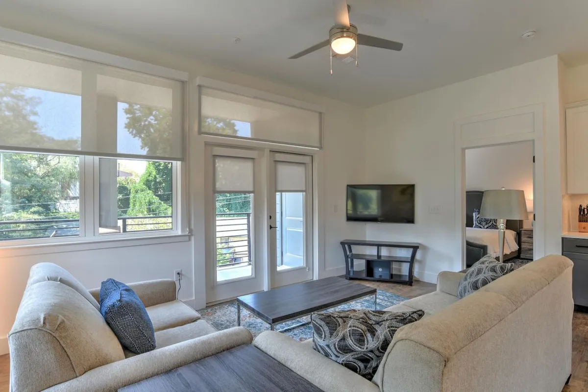 A cozy living area featuring a comfortable sofa, coffee table, large windows that allow plenty of natural light, and a television. The view through the windows suggests a connection to the outside world, helping guests unwind after a busy day in downtown Asheville.