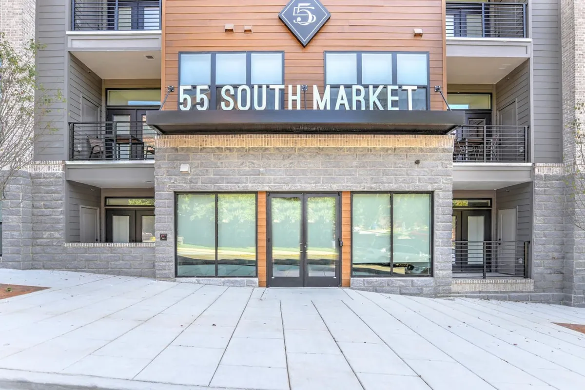 The image features the exterior of a modern building with large glass windows and prominent signage. The building faces a welcoming street with patio space, inviting pedestrians and residents to enjoy the vibrant neighborhood. The scene exudes a sense of community engagement and urban excitement.