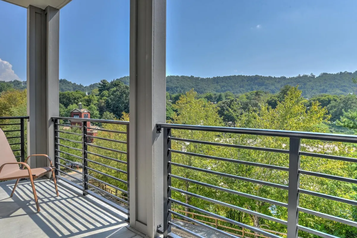 The hero image features the condo's balcony overlooking lush green trees and gentle hills, with soft morning light suggesting a tranquil sunrise. The sturdy balcony railing invites guests to step outside and take in the views, creating a serene spot for morning coffee or evening stargazing. The peaceful scene is framed by nature, with hints of wildlife and the appeal of a private outdoor space just steps from downtown Asheville.